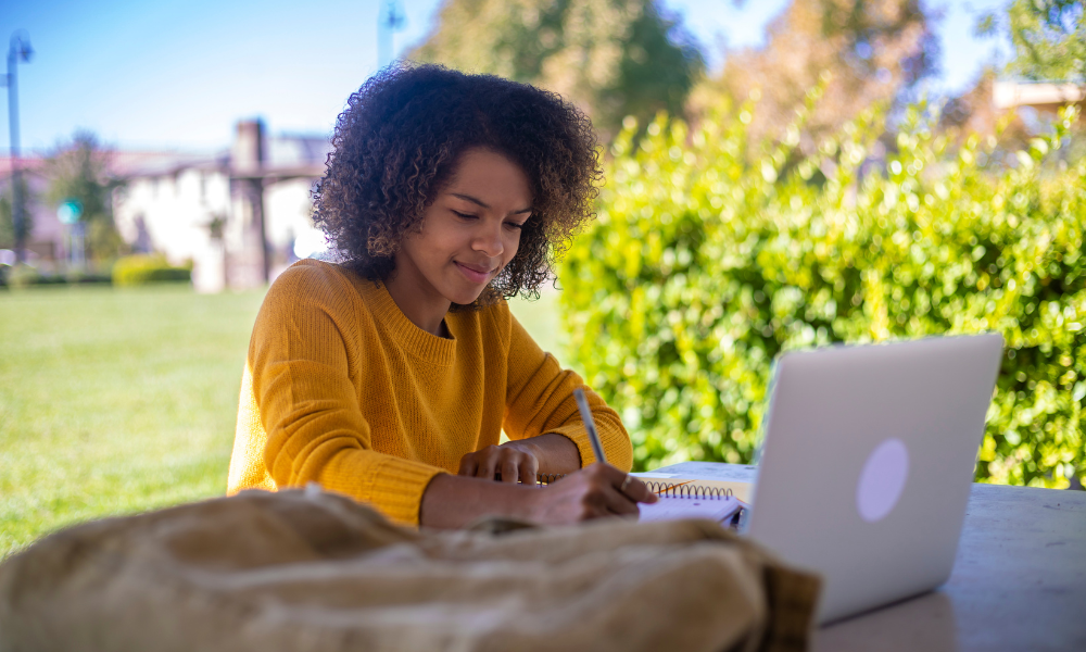 A college student sitting outside of the library and she is applying for May scholarships on her laptop, higher education, high school students, undergraduate students, graduate students, minority groups, scholarships with upcoming deadlines, end of may scholarships,