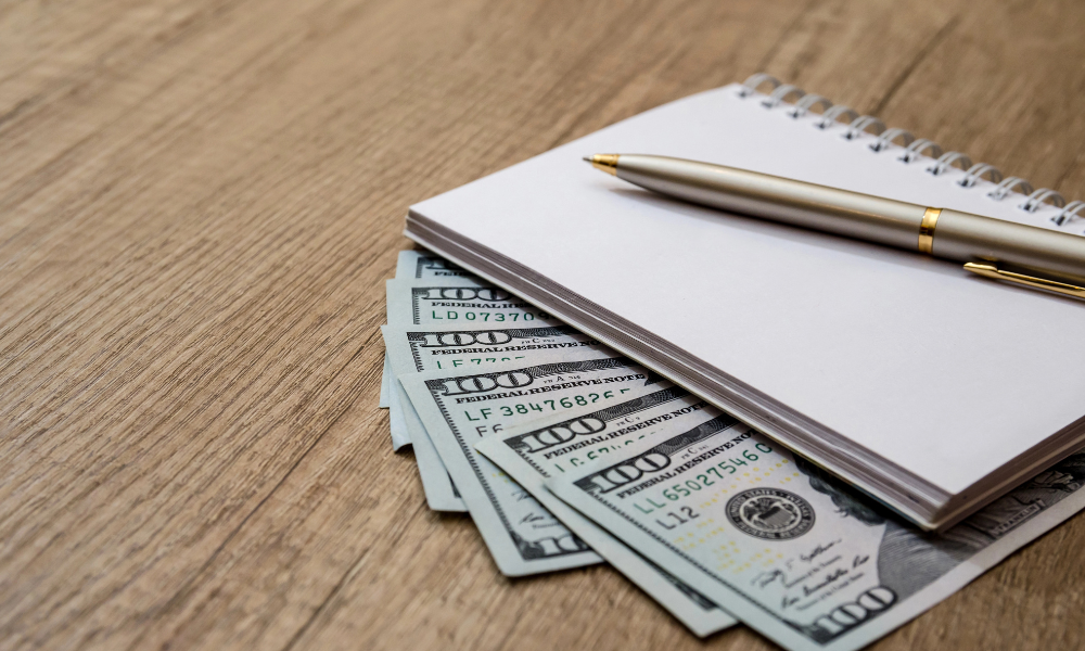A wad of hundred dollar bills with a notebook and a pen on top, scholarship money, scholarship winner, scholarships with January deadlines, scholarships due in late January
