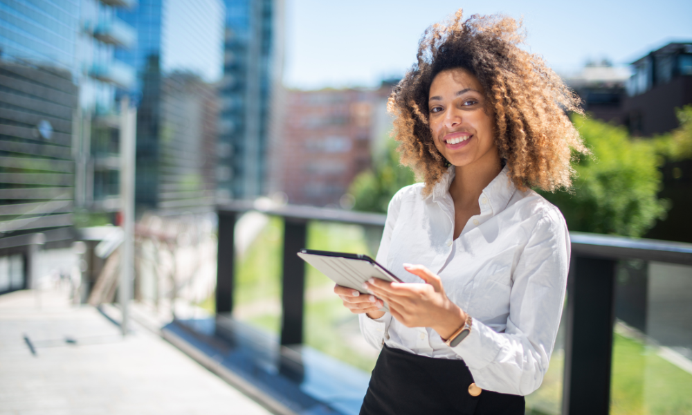 A business student is standing outside of the business school looking on her laptop for business scholarships for graduate students, MBA students, current college students, college bound seniors, high schoolers, high school seniors, general business, international business majors, information technology majors, entrepreneurship majors