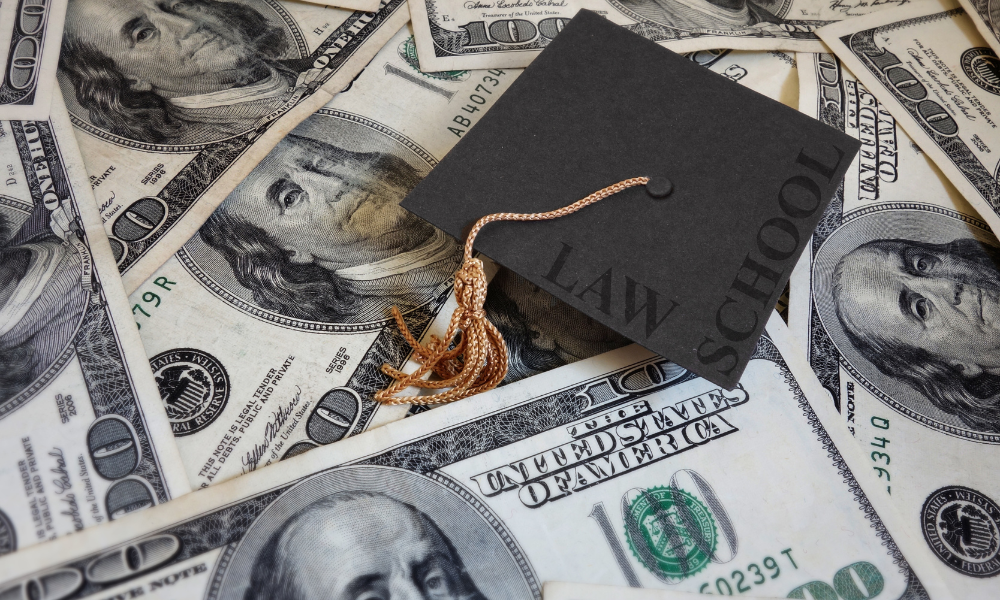 A law school graduation cap is sitting on top of a pile of scholarship money a student won from applying for law scholarships with urgent summer deadlines. The student also applied to full-ride scholarships, merit scholarships, and first generation scholarships